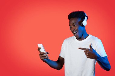 Neşeli mutlu genç zenci adam müzik dinliyor ya da internetten görüntülü içerik izliyor, akıllı telefon ve kablosuz kulaklık kullanıyor, elindeki cep telefonunu işaret ediyor, kırmızı stüdyo arka planı, kopyalama alanı.