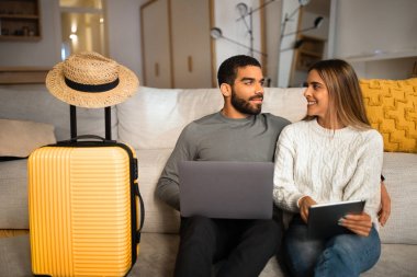Neşeli uluslararası milenyum ailesi oturma odasında tatil için ellerinde cihazlar, bavullar ve şapkalarla yerde oturuyorlar. Evde tatil için hazırlan, seyahat planı, seyahat uygulaması