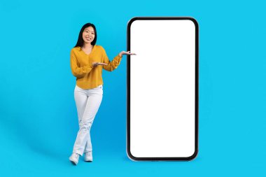 Çekici, neşeli, genç Asyalı bir kadın beyaz boş cep telefonuyla güzel mobil uygulamalar gösteriyor. Mavi stüdyo arka planı, maket, kopyalama alanı. Online teklif, çok iyi.