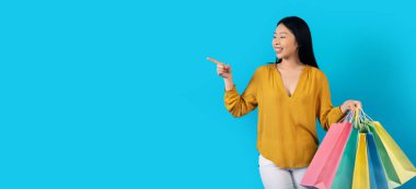 Neşeli, çekici, genç Koreli kadın renkli alışveriş torbaları taşıyor mavi stüdyo arka planında fotokopi alanını işaret ediyor, web-afişi. Perakende satış, tüketim, alışveriş