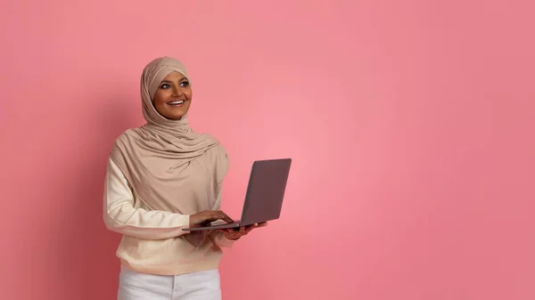 Pensive Young Muslim Woman With Laptop In Hands Standing Over Pink Studio Background, Smiling Islamic Lady Wearing Headscarf Holding Computer, Enjoying Online Job Opportunities And Remote Work