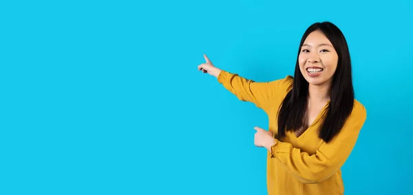 Laughing beautiful millennial chinese lady in nice outfit posing on blue background, pointing at copy space behind her back and smiling, young asian woman showing nice offer, panorama