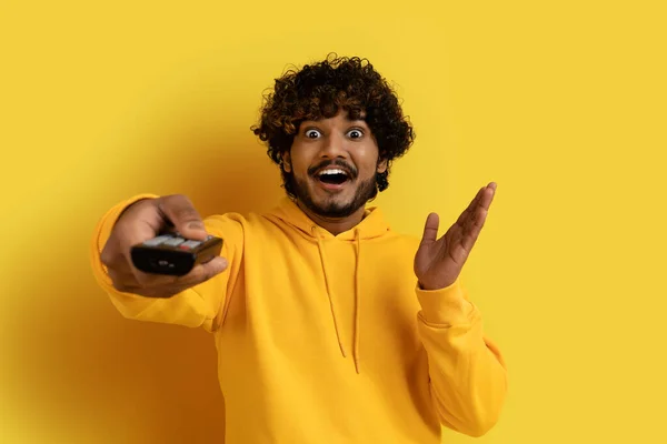 快乐的情绪英俊的卷曲的印度年轻人 身穿黄色的帽衫 手持电视遥控器 在黄色的工作室背景上做手势 欣赏电视节目 复制空间 — 图库照片