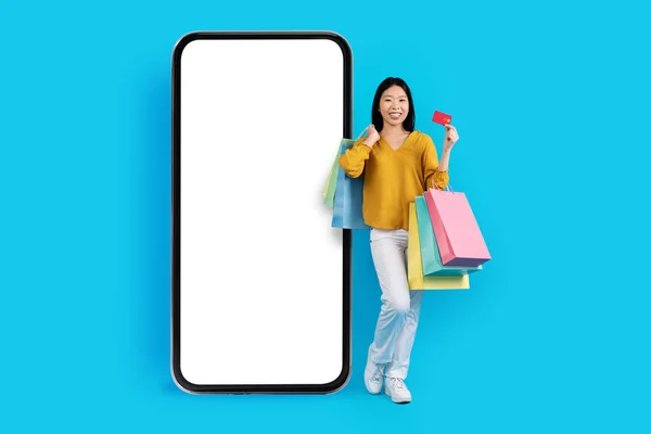 积极快乐的笑着年轻的亚洲女士 带着五颜六色的购物袋和银行卡 摆设在白色空白屏幕的大手机旁边 女性推荐网上购物 — 图库照片