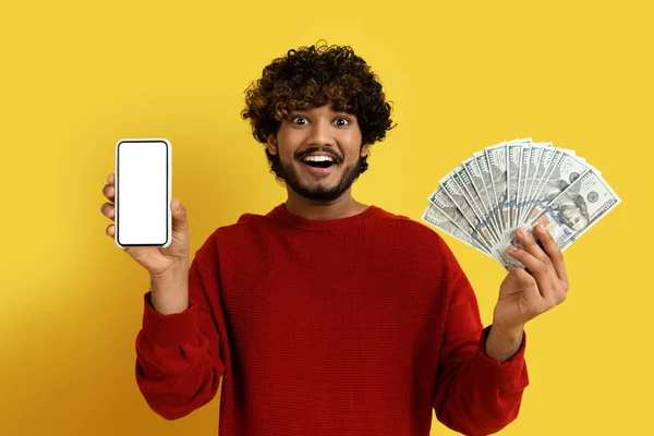 在线贷款 快乐英俊的印度年轻人 穿着红色毛衣 拿着现代手机 屏幕是白的 钞票是钱做的 背景是黄的 是个模特儿 — 图库照片