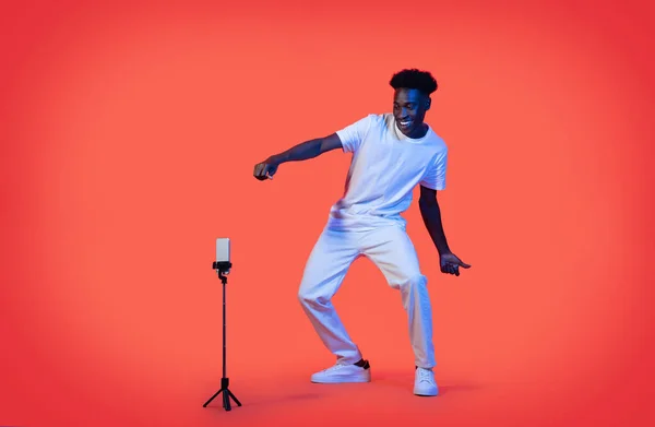 有名なインフルエンサークールなスタイリッシュなミレニアルブラック男は ソーシャルメディアのためのコンテンツを記録し 三脚に設定された携帯電話を見て ネオンの光の中で赤の背景に踊りと笑顔 完全な長さ — ストック写真