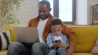 Modern ailelerde Gadgets bağımlılığı sorunu. Genç Afrikalı Amerikalı baba ve küçük oğlunun laptopunu ve akıllı telefonunu kullanarak evde ağır çekimde otururken çekilmiş bir portresini izliyoruz.
