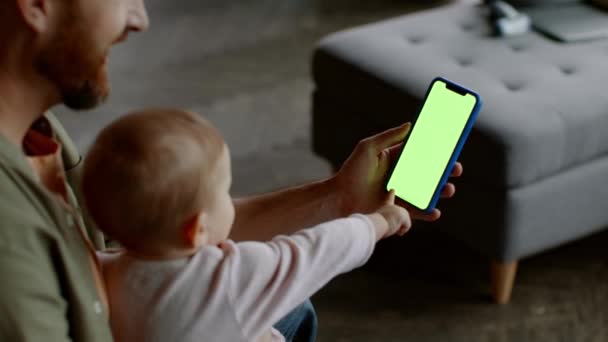 若い男のウェブサーフィンスマートフォンで緑のクロマキー画面 好奇心小さな赤ちゃんは ガジェットに触れようとしています 肩のビューの上 スローモーション 空のスペース — ストック動画