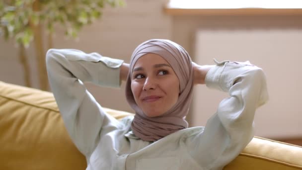 休息和幸福 中东无忧无虑的年轻女子的近照 头戴头巾 在家里放松下来 双手靠在沙发上笑着 跟踪镜头 慢动作 自由自在 — 图库视频影像