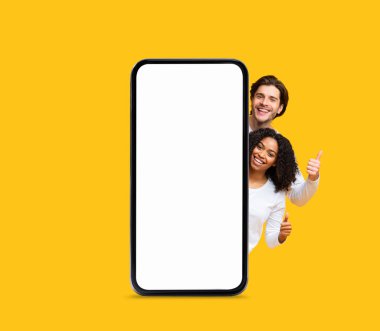 Güzel teklif. Mutlu Çoklu Etnik Çift, Büyük Boş Akıllı Telefonun Arkasına Gizlice Bakıyor ve Başparmaklarını Sarı Arkaplanda Gösteriyor Neşeli Erkek ve Kadın Yeni Mobil Uygulama veya Çevrimiçi Teklif Ediyor,