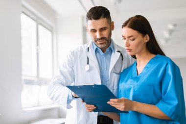 Orta yaşlı erkek pratisyen doktor ve genç bayan asistan hastanede görevleri tartışıyor, kadın ve erkek panoda bilgi arıyor, boş alan