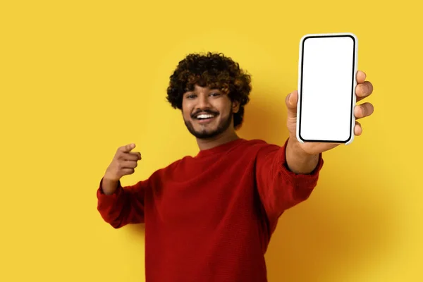 笑一个英俊时髦的印度年轻人 带着白色空白屏幕在镜头前展示着巨大的智能手机 模仿在线提供或移动应用程序广告 被黄色工作室背景隔离 复制空间 — 图库照片