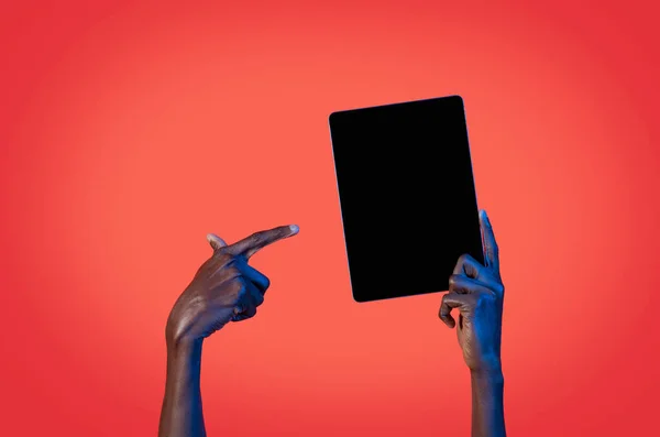 Unrecognizable black man pointing at modern digital tablet with black empty screen over red studio background in neon light, cropped of african guy recommending newest mobile app, mockup