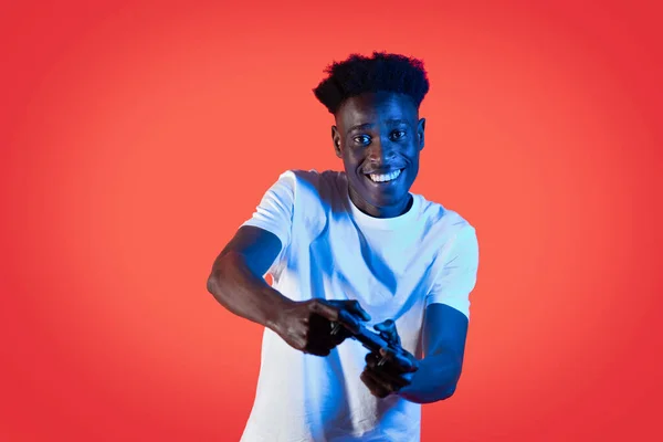 Addiction, gaming disorder concepy. Happy cheerful young black guy in white t-shirt playing video game, holding joystick console and smiling, red studio background with neon light, copy space