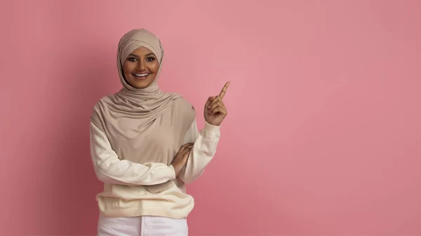 看看这个快乐的穆斯林女性在Hijab中指指点点在粉红背景之上的复印空间 在头巾中微笑的伊斯兰女性展示你的广告的自由位置 — 图库照片