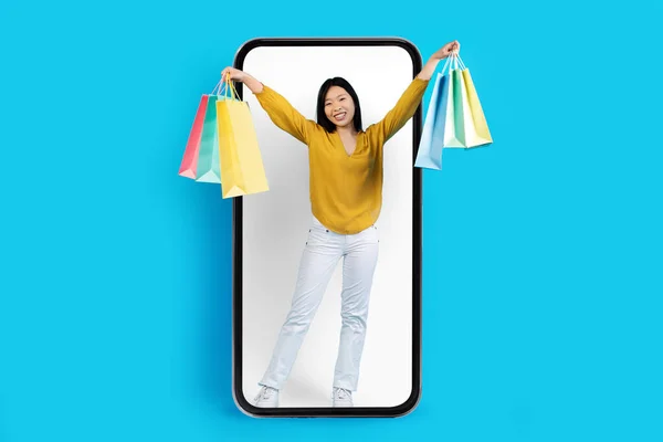 网上购物 电子商务 快乐而美丽的亚洲年轻女子举手投足 提着五颜六色的购物袋 站在装有白色屏幕和蓝色工作室背景的大型智能手机里 — 图库照片