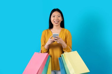 Bin yıllık neşeli gülümseyen, günlük kıyafet alışverişi yapan, elinde renkli kağıt torbalarla gezen, cep telefonu kullanan, gülümseyen, mavi arka planda izole edilmiş Çinli kadın. İnternetten alışveriş