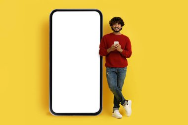 Havalı, şık, genç doğulu hippi beyaz ekranlı akıllı telefonun yanında poz veriyor, cep telefonu kullanıyor, güzel mobil uygulama kullanıyor, sarı Stuido arka planında model alıyor, tam boy.