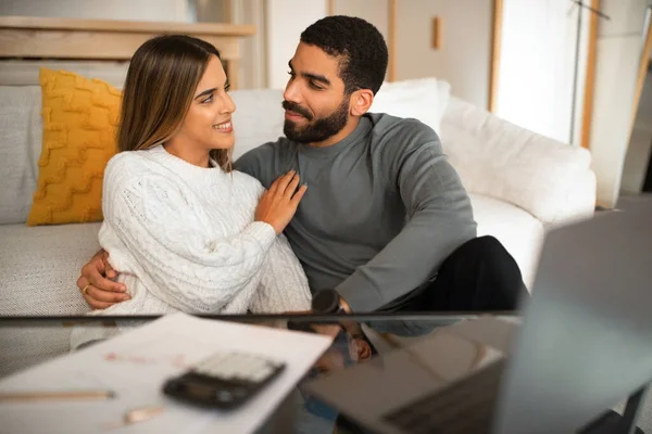 Happy international millennial couple hugging, use laptop, have video call, pay taxes in living room interior. Love and relationship, bookkeeping and finance together at home