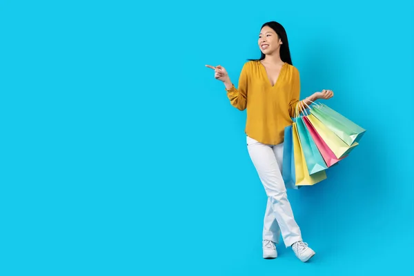 穿着休闲装 笑容可亲的 有魅力的 长发的年轻的大韩民国女士 带着五颜六色的购物袋 在蓝色的工作室背景上展示出广告的自由空间 — 图库照片