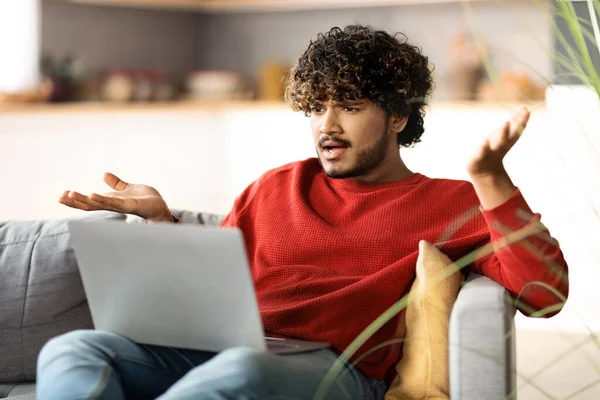 Stressed Young Indian Man Suffering Problems While Using Laptop At Home, Anxious Eastern Guy Looking At Computer Screen And Emotionally Gesturing While Sitting On Couch In Living Room, Closeup