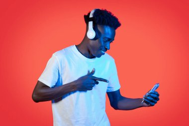 Gıcır gıcır saçlı, yeni kablosuz kulaklık takan ve elinde akıllı telefonuyla kırmızı arka plandaki neon ışığı altında Afro-Amerikan bir adamın müzik uygulamasından hoşlanması.