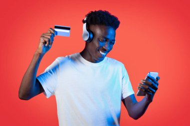 E-ticaret, e-bankacılık. Neşeli, mutlu, beyaz tişörtlü genç adam akıllı telefon, kulaklık ve banka kartı kullanıyor, mallar için ödeme yapıyor, kırmızı stüdyo ışıklarıyla internet üzerinden servis yapıyor.