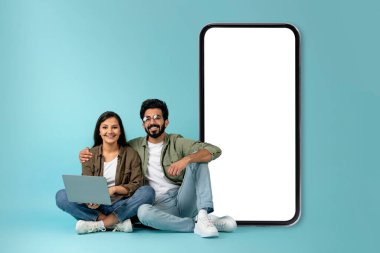 Neşeli, neşeli, milenyum melez bir çift mavi arka planda rahat poz veriyor, modern bilgisayar kullanıyor, beyaz ekranlı büyük bir telefonun başında oturuyor, maket yapıyor, uzay kopyalıyor.
