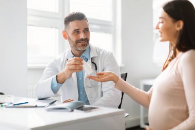 Jinekoloji, tıp ve hamilelik kavramı. Erkek jinekolog, hastanedeki randevusu sırasında gülümseyen hamile kadına hap veriyor. Doktor konseptini ziyaret et
