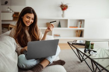 Gülümseyen milenyum melez kadın dizüstü bilgisayarda alışveriş yapıyor, kredi kartı kullanıyor, oturma odasında online alışveriş yapıyor. Uzaktan iş, satış, bankacılık ve nakit para evde
