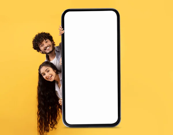 不错的提议 快乐的印度夫妇站在大大的空白智能手机后面 白色的屏幕笼罩在黄色的背景下 快乐的年轻人和女人在演示应用程序或网站设计的复制空间 — 图库照片