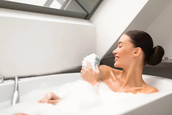 現代の浴室の屋内で目を閉じて泡のポーズでお風呂を取るスポンジできれいな女性の洗濯ボディ 浴槽に寝そべっている女 ボディケアとスパのコンセプト 選択的フォーカス — ストック写真