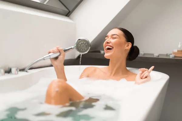 快乐的女士 一边洗澡一边唱歌一边抱着淋浴头 就像麦克风一样 坐在水里 在家里的现代浴室里 泡沫泡在水里 美感与放松 — 图库照片