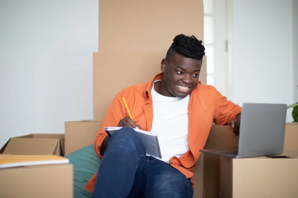 Happy Black Man Making Checklist Necessities Using Laptop Moving Home — Φωτογραφία Αρχείου