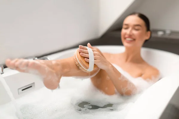 泡でお風呂に入る木製のブラシで豪華な女性洗濯マッサージボディバスルームの屋内で滑らかな脚をこすります ボディケアとスキンケア化粧品のコンセプト 選択的フォーカス — ストック写真