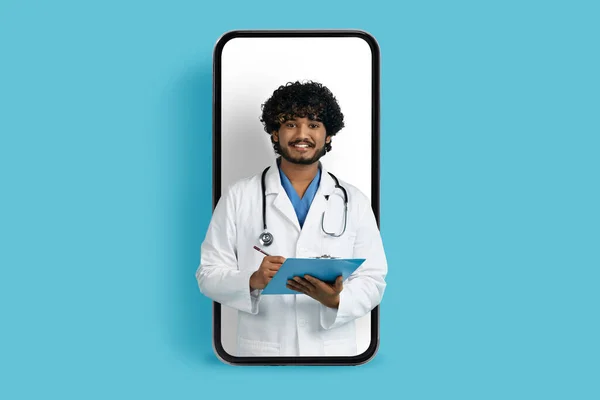 Handsome cheerful young indian man in in white medical coat doctor have online meeting with patient, male family doc posing in huge cell phone screen, collage, blue studio background