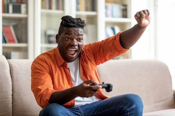 在家里休息 家庭娱乐的概念 动情的年轻非洲裔美国人 头戴辫子 玩电子游戏 举手大笑 一个人坐在家里沙发上 抄袭空间 — 图库照片