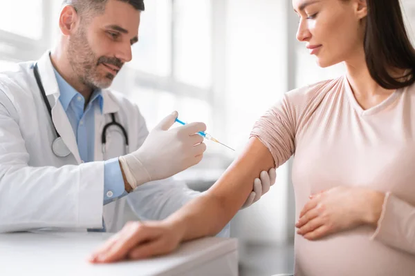 Vaccination in pregnancy. Young pregnant woman getting vaccinated at hospital, male doctor giving vaccine shot injecting lady in arm. Covid-19 and flu prevention