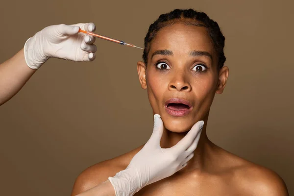 Shocked black middle aged woman receiving beauty shots, using injection medicine to prevent aging, brown studio background. Aged woman having injections of collagen or hyaluronic acid