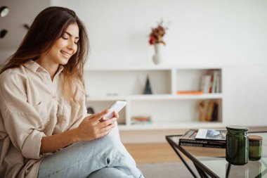 Milenyum melezi gibi gülümseyen bir kadın telefona bakıyor, mesaj okuyor, sohbet ediyor, internette oda içinde sörf yapıyor. Uzaktan iletişim uygulaması, sosyal ağlar, evde iyi haberler