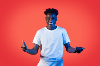 Beyaz giyimli mutlu yakışıklı genç adam neon ışıkta kırmızı arka planda izole edilmiş, neon ışıkları altında neşeli ve gösterişli tutarak şampiyon oluyor, modern eğlence teknolojileri, video oyunları