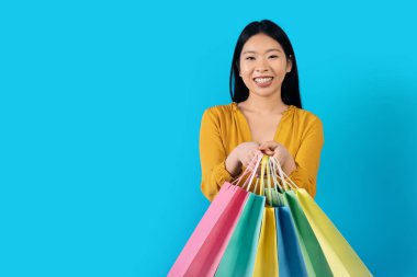 Renkli alışveriş torbaları tutan, kameraya gülümseyen, mavi stüdyo arka planında izole edilmiş, sarı elbiseli, güzel Asyalı bir kadın. Sezon indirimi, Kara Cuma konsepti.