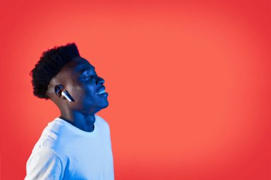 Neşeli, güler yüzlü, şık, beyaz tişörtlü Afro-Amerikan adam. Yeni kablosuz kulaklıklarla müzik dinliyor. Kırmızı kulaklıklar, neon ışığı, fotokopi odası, yan görüş.