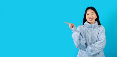 Neşeli gülümseyen mavi örgü süveterli güzel Koreli kadın mavi stüdyo arka planında reklam için boş yer gösteriyor, güzel bir anlaşma öneriyor, panorama, afiş.