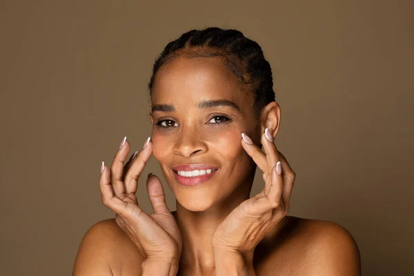 Beauty face. Happy black middle aged woman touching healthy skin and smiling, female with fresh glowing hydrated facial skin and natural makeup on brown background