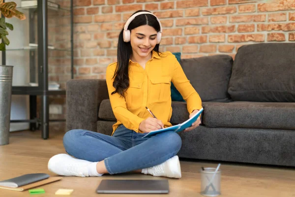 Cheerful spanish student lady in wireless headphones taking notes, watching video lesson on laptop in living room interior. Study at home alone, knowledge and webinar with device