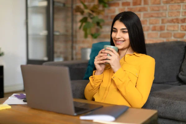 家でコーヒーを飲みながら ノートパソコンを使って 画面を見て 笑顔で 映画を見て 休憩しながら インターネット上でビデオを見ている正のスペイン人女性 — ストック写真