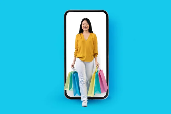 穿着休闲装的年轻亚洲女人带着购物袋和蓝色的工作室背景 带着白色屏风走出大手机 网上购物 电子商务 移动应用 — 图库照片
