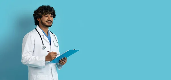 穿着工作服 面带微笑 英俊而开朗的印度年轻医生 带着听诊器和医学图表 在蓝色的工作室背景上看着复制的空间 网页横幅 医疗概念 — 图库照片