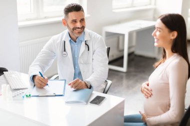 Doğum öncesi bakım konsepti. Klinikte oturan, erkek jinekologla konsolosluk yapan, göbeğini kucaklayan ve doğum uzmanıyla konuşan mutlu, genç hamile bir bayan.